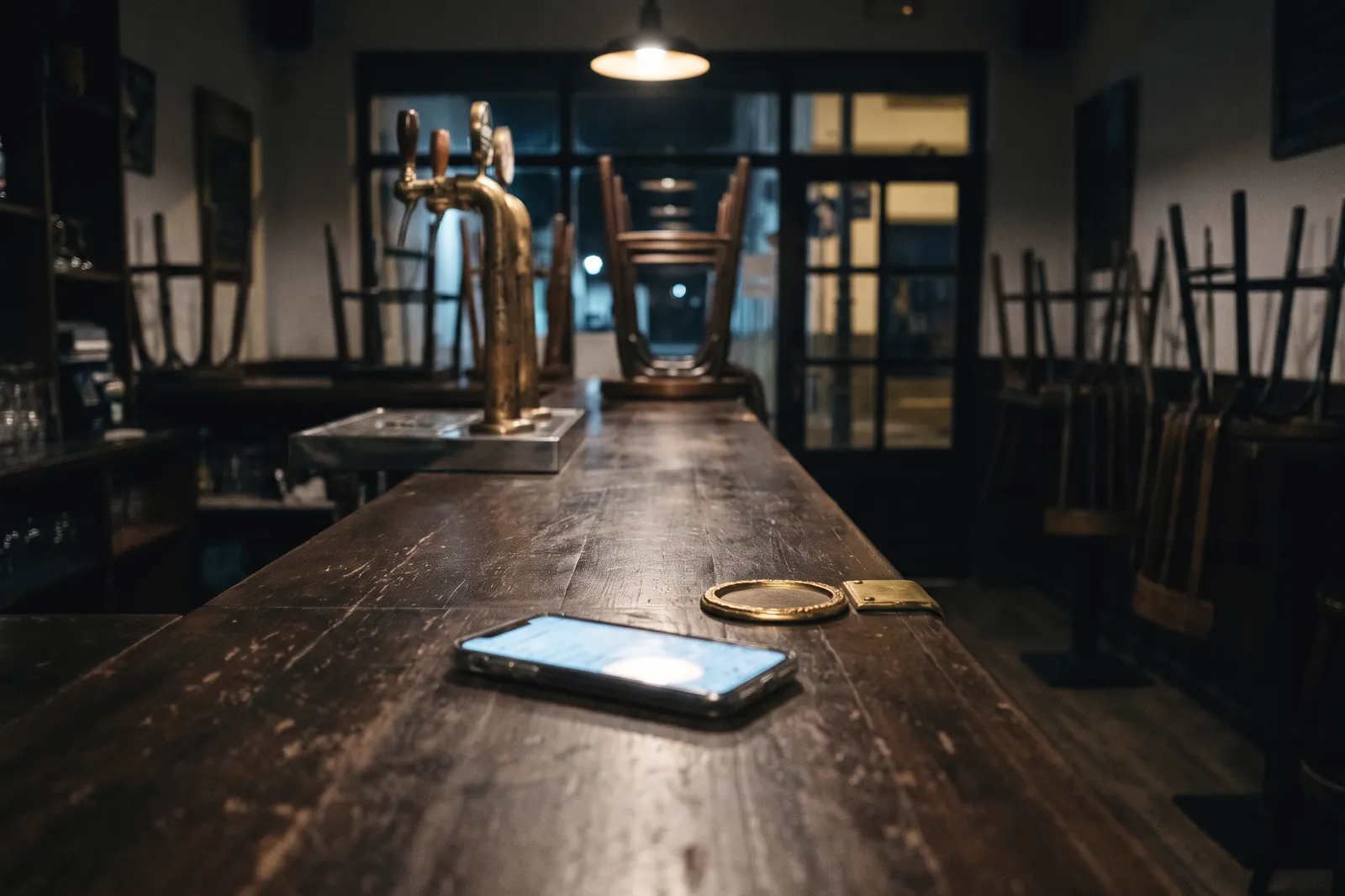 Barra de madera de un bar ya cerrado, de noche, con un móvil encendido encima y las sillas subidas