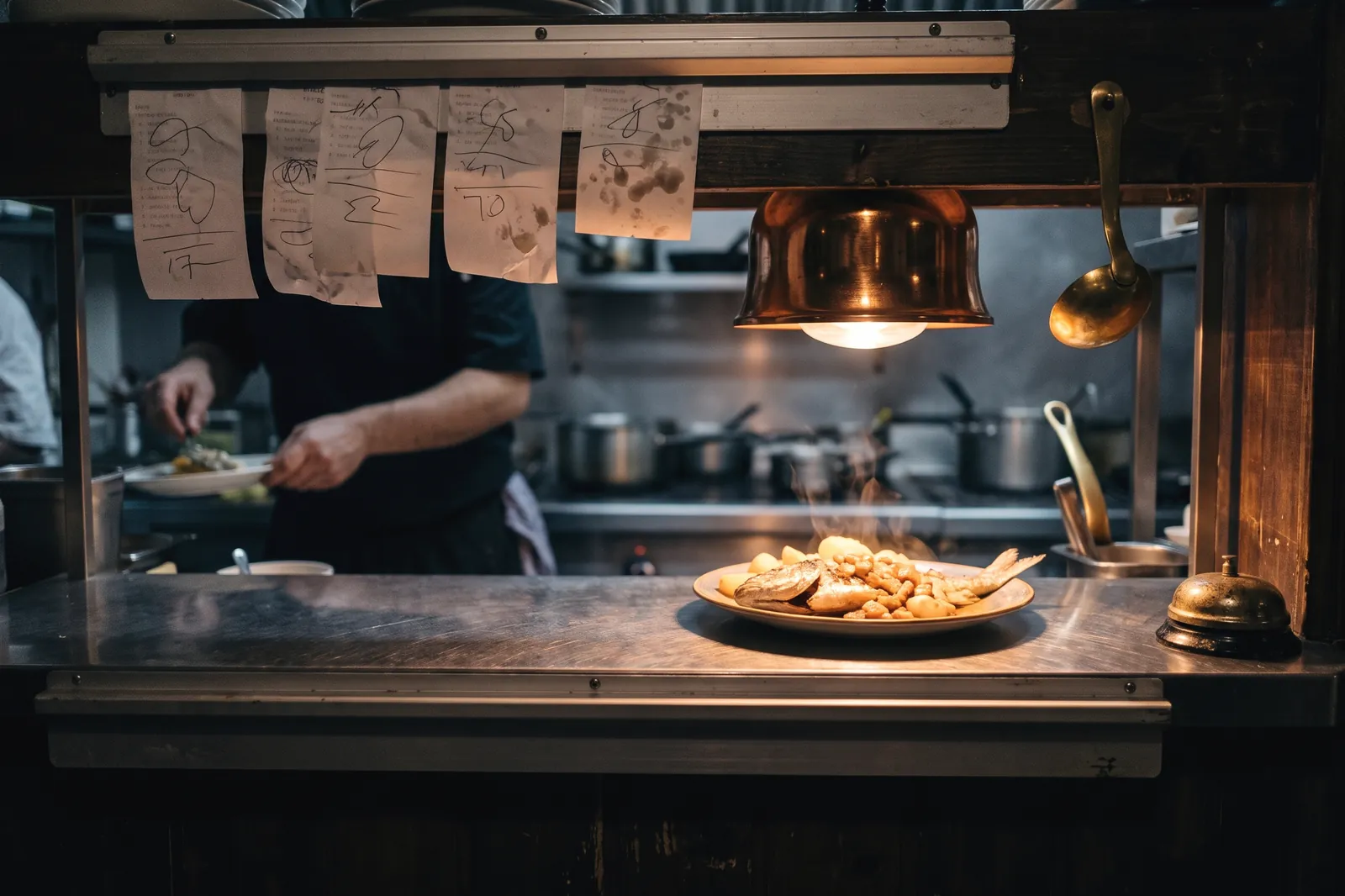 Pase de cocina de un restaurante en pleno servicio, con comandas colgadas y un plato de pescado bajo la lámpara